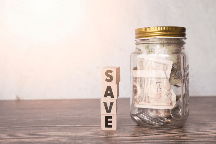 Glass jar filled with coins and paper currency next to wooden blocks spelling "SAVE," symbolizing the concept of saving money on a wooden surface with a neutral background.