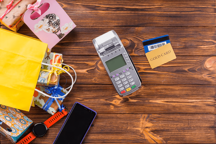 A credit card payment terminal surrounded by a gold card, shopping bags, gift boxes, smartwatch, and smartphone on a wooden table, representing modern retail transactions and digital payments.