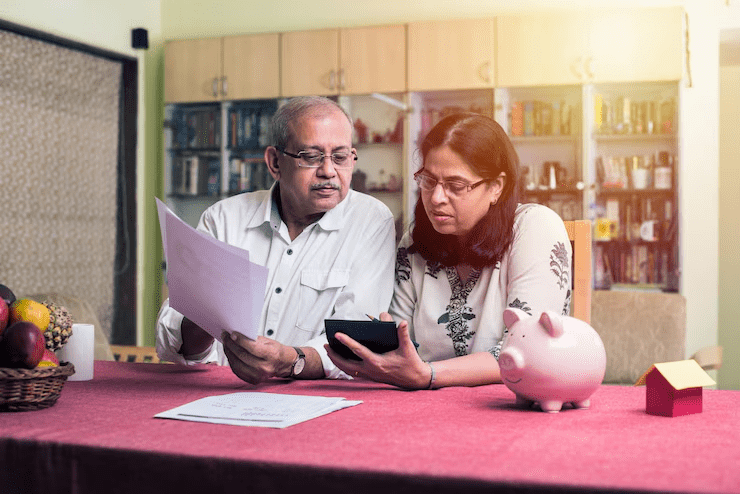 "Senior couple sitting at a table reviewing financial documents together, with the woman using a calculator and the man holding papers; a piggy bank and miniature house model are also on the table, symbolizing financial planning or budgeting."