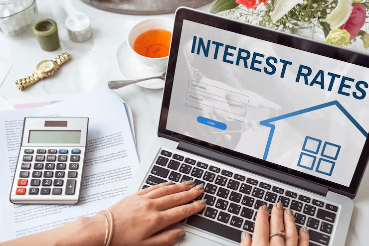 "Person typing on a laptop displaying a login screen with the title 'Interest Rates', alongside documents, a calculator, a cup of tea, and flowers on the table, indicating financial research or mortgage planning."
