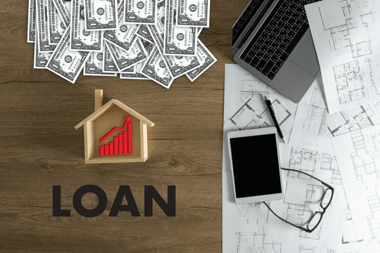 "Top view of a desk with scattered dollar bills, a miniature wooden house with an upward red graph, the word 'LOAN', architectural blueprints, a laptop, tablet, pen, and eyeglasses—symbolizing home loans or real estate financing."
