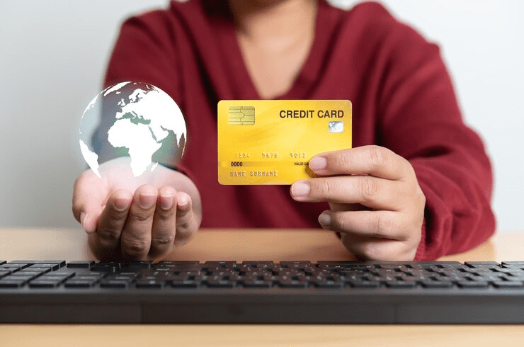 "Person sitting at a desk holding a yellow credit card in one hand and a glowing digital globe in the other, with a keyboard in front, symbolizing global online payments or international e-commerce."