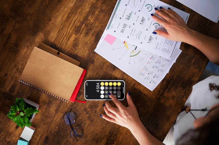 This images shows a Alt Text: Financial planning concept with stacked coins, model house, car, and US dollar bills on a desk, with a businessman using a smartphone in the background