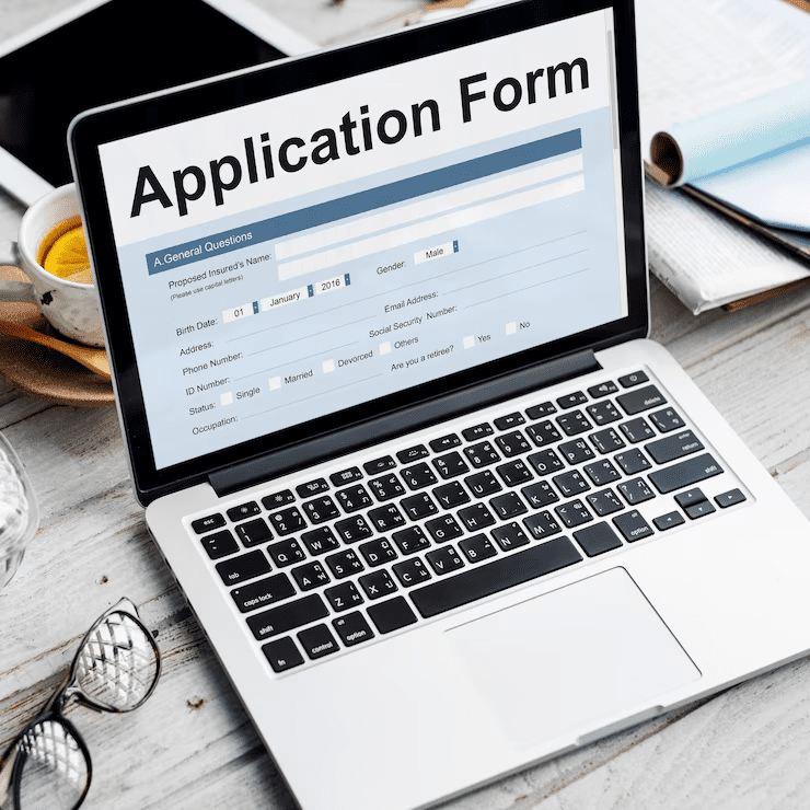 Laptop displaying an online application form on screen, placed on a desk with documents, glasses, and a cup, representing the digital application process for Canara Bank gold loan.