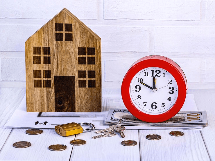 Wooden house model beside a red alarm clock, dollar bills, padlock with keys, and scattered coins, representing real estate, mortgage timing, and financial security.