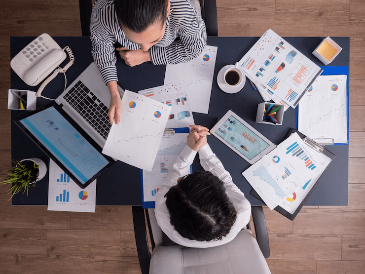 Top-down view of two professionals analyzing financial charts and graphs at a desk with a laptop, tablet, printed reports, coffee cup, and office supplies.