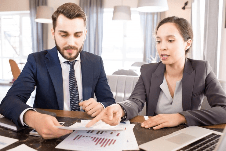 A businesswoman and businessman in formal attire reviewing financial documents with charts at a desk, engaged in a professional discussion.