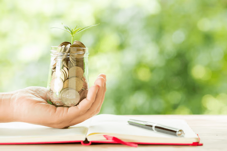 A hand holding a glass jar filled with coins and a small green plant sprouting from it, placed over an open notebook with a pen, symbolizing savings and financial growth.
