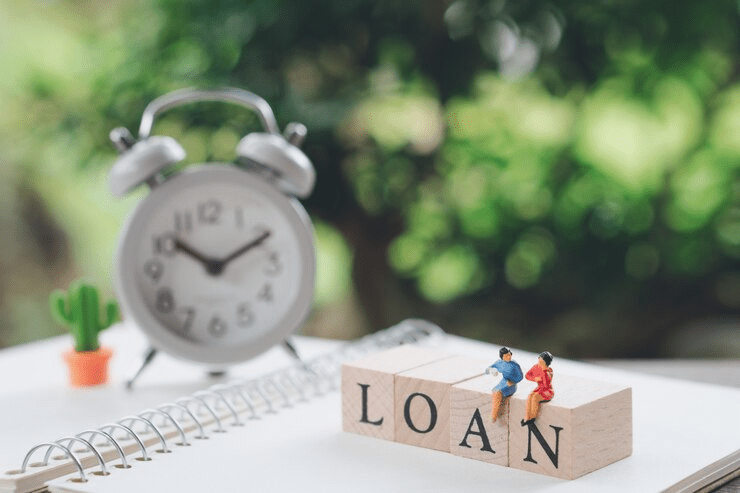 Miniature figurines sitting on wooden blocks spelling “LOAN,” placed on a notebook beside a classic alarm clock and a small cactus, symbolizing loan timing and financial planning.