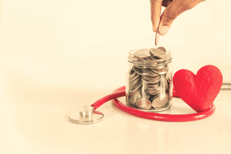 A hand placing a coin into a glass jar filled with coins, surrounded by a red stethoscope and a red heart, symbolizing health savings, medical funds, or health insurance planning.