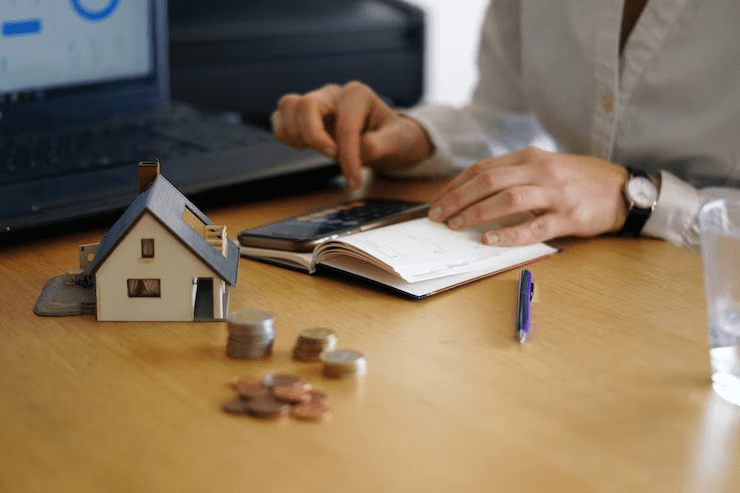 A person using a calculator and writing in a notebook on a desk with a miniature house model, coins, a pen, a laptop, and a glass of water—symbolizing home loan planning, budgeting, or mortgage calculation.
