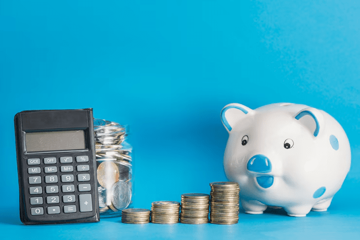 A black calculator, a glass jar filled with coins, stacked coins arranged in ascending order, and a white piggy bank with blue spots, all set against a blue background—symbolizing budgeting, saving, and financial planning.