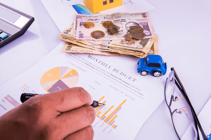 A hand holding a pen pointing at a bar chart on a monthly budget sheet, surrounded by Indian currency notes and coins, a toy blue car, glasses, and a calculator—symbolizing financial planning, budgeting, and expense tracking.