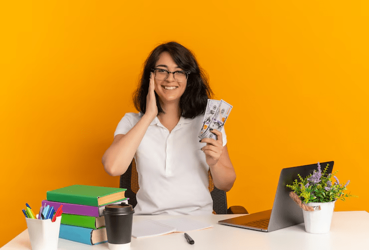 A smiling woman sitting at a desk holding a fan of U.S. dollar bills, surrounded by books, a coffee cup, stationery, a laptop, and a small potted plant—symbolizing financial success, payday loans, or student funding.