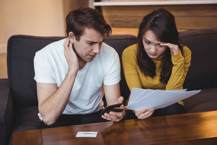 A worried young couple sitting on a couch, looking at financial documents and a smartphone, appearing stressed about bills or finances.