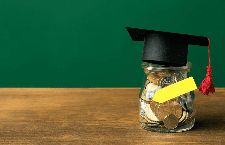 Glass jar filled with coins wearing a graduation cap, placed on a wooden table against a green background — representing education savings or student loans.