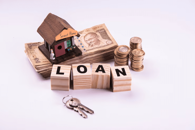 Wooden blocks spelling "LOAN" placed in front of a miniature house labeled "Sweet Home," with Indian currency notes, coin stacks, and house keys, symbolizing a home loan or mortgage concept.