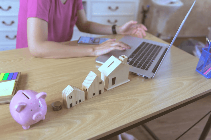 Person working on a laptop at a desk with a pink piggy bank, coins, and miniature wooden house models, symbolizing financial planning and home savings.








