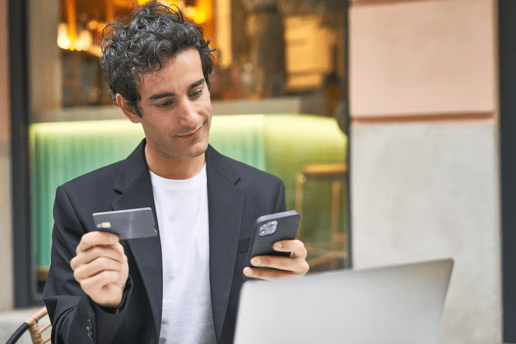Man holding a debit or credit card in one hand and a smartphone in the other while sitting in front of a laptop, representing the eligibility and requirements for NEFT transactions through digital banking platforms.