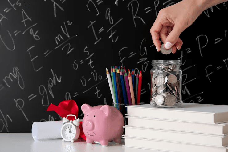 Hand placing a coin into a glass jar filled with coins, surrounded by a pink piggy bank, stack of books, colored pencils, a diploma scroll with a ribbon, and an alarm clock, set against a blackboard with chalk mathematical formulas—symbolizing saving for education.