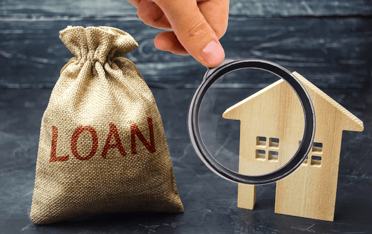 A hand holding a magnifying glass near a burlap sack labeled "LOAN" and a wooden model house, symbolizing home loan scrutiny or financial analysis.