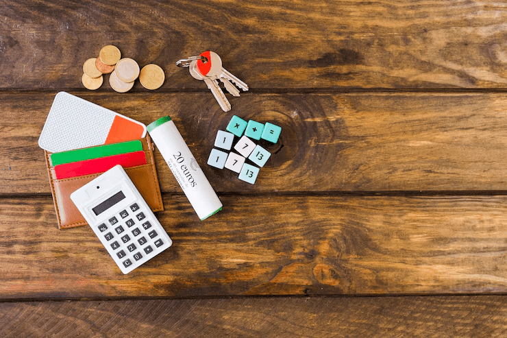 Assorted financial and educational items on a wooden surface, including a calculator, coins, wallet, keys, dice with numbers and math symbols, and a roll labeled "20 CURSOS," representing budgeting or academic planning.
