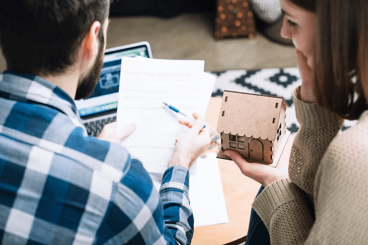 "Couple reviewing documents and holding a small wooden house model, representing home loan application or property purchase planning."

