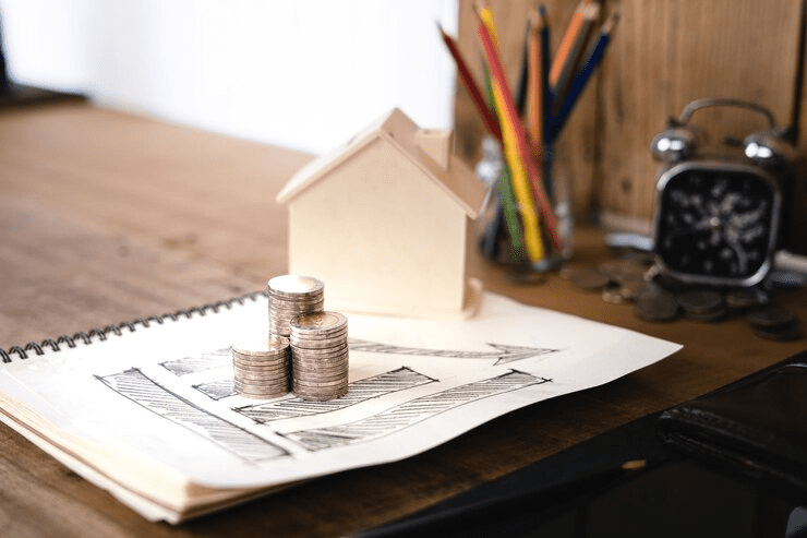 The image shows a small, unpainted wooden model of a house placed on a notepad with hand-drawn upward trending arrows. In front of the house are three stacks of coins of varying heights, suggesting growth. The setting appears to be a desk with a jar of colored pencils and an alarm clock in the background. The overall composition conveys the concept of increasing investment or savings related to home ownership