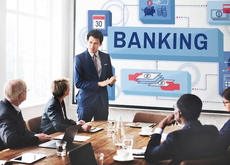 Business professionals in a meeting room with a presenter explaining banking concepts in front of a large screen displaying the word “BANKING” along with financial icons, representing corporate banking strategy or financial planning discussion.