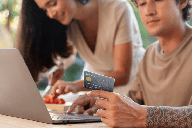 Man using a laptop while holding a credit card, with a woman chopping vegetables in the background, symbolizing online shopping, digital payments, or managing finances from home in a casual setting.