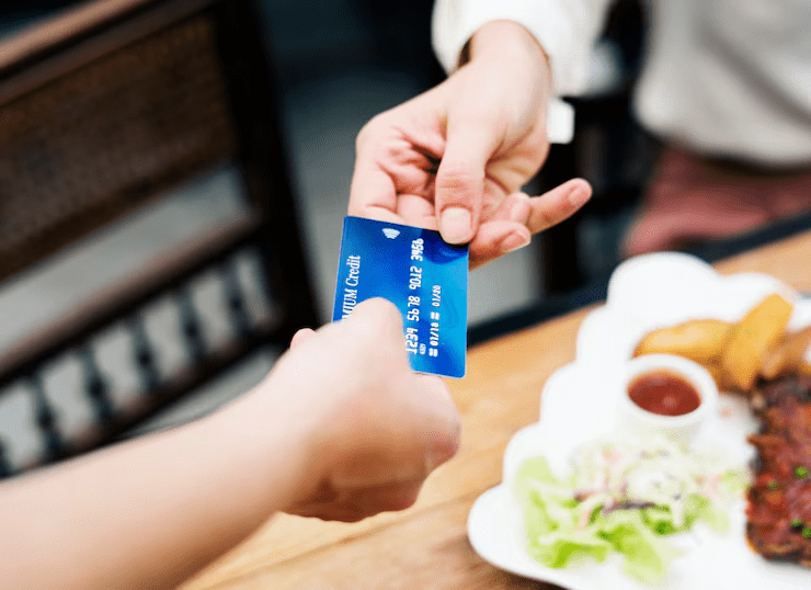 Close-up of a person handing over a blue credit card for payment at a restaurant, with a plate of food in the background—representing credit card usage for dining and cashless transactions.