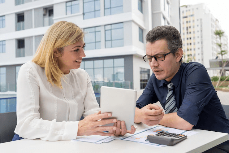 Two professionals discussing financial documents and analyzing data on a tablet during a financial planning consultation outdoors, with buildings in the background.