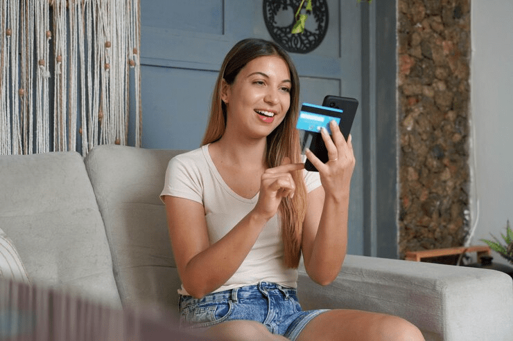 Young woman sitting on a couch using a smartphone with a credit card in hand, making a transaction via a credit card payment app.